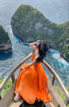 a woman in an orange dress is sitting on a bench looking out at the ocean