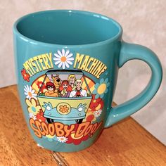 a blue coffee mug with the words mystery machine and cartoon characters on it sitting on a wooden table