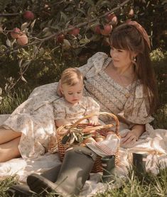 a woman and child sitting in the grass under an apple tree