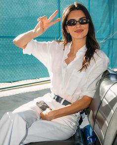 a woman sitting on top of a car wearing sunglasses and making the peace sign with her hand