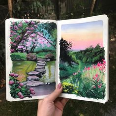 someone is holding up an open book with watercolors on it and trees in the background