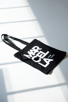 A canvas bucket tote bag in 100% European cotton, with a bottom panel for added capacity. The tote features an oversized MARCELLA NEW YORK graphic on one side, in white print on black canvas.[SPLIT] Our production team enjoys a living wage, a 40-hour work week with paid overtime, free health insurance and 20 days annual paid vacation. White Canvas Bag With Logo, Cotton Tote Shoulder Bag With Logo Print, Canvas Tote Shoulder Bag With Logo Print, Travel Cotton Shoulder Bag With Logo Print, Everyday Cotton Bag With Logo, Travel Shoulder Bag With Logo Print In Cotton, Everyday Cotton Shoulder Bag With Logo, Daily Use Cotton Bag With Logo, Everyday Cotton Shoulder Bag With Logo Print