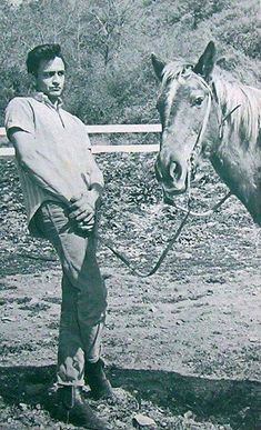 a man standing next to a horse on a dirt field with trees in the background