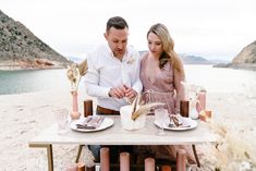 a man and woman sitting at a table on the beach with desert scenery in the background