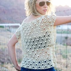 a woman leaning against a fence with her arms behind her back, wearing sunglasses and a crocheted top