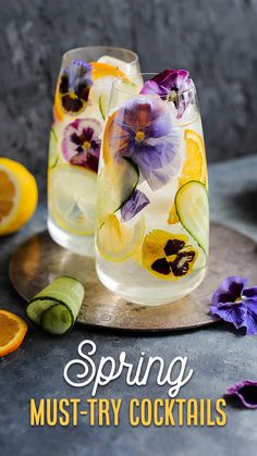 two glasses filled with flowers and cucumbers next to sliced oranges on a tray