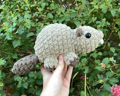 a hand holding a small stuffed animal in front of some bushes and flowers with green leaves