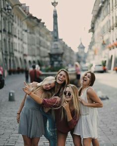 three girls are posing for the camera in front of some buildings and one girl is holding her hand up