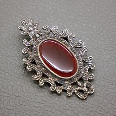 This exquisite vintage brooch is a true statement piece, featuring a striking brown-red carnelian gemstone set in sterling silver. Crafted circa 1950s -1970s, this brooch showcases a timeless Edwardian style adorned with sparkling marcasites. The intricate silver work beautifully complements the smooth, polished carnelian, making it a perfect addition to any vintage jewelry collection. Whether worn on a jacket lapel, dress, or scarf, this large silver brooch adds a touch of elegance and sophisti Vintage Gemstone Brooches For Formal Occasions, Vintage Silver Brooch With Gemstone, Vintage Silver Gemstone Brooch, Ornate Red Jewelry Brooch, Vintage Gemstone Brooches, Ornate Gemstone Brooches For Collectors, Elegant Red Brooches Collectible, Ornate Cabochon Brooch For Formal Occasions, Ornate Cabochon Brooches For Formal Occasions