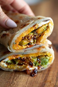 a person holding a burrito in their hand on a wooden table with other food items