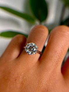 a woman's hand with a diamond ring on top of her finger and a plant in the background