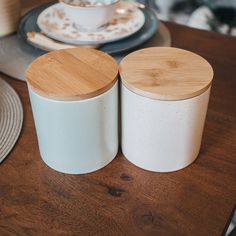 two white containers sitting on top of a wooden table next to a plate and cup