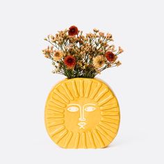 a yellow vase with flowers in it and a face on the front, against a white background