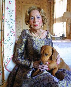 an older woman sitting on a bed holding a dog in her lap and looking at the camera