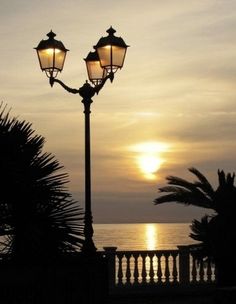 the sun is setting over the ocean and some lights are lit up in front of an iron fence