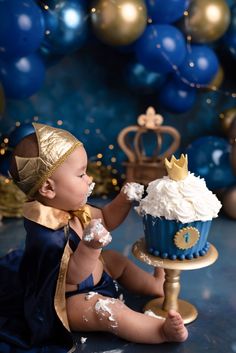 a baby is sitting on the floor with a cupcake in front of him and blue balloons behind him