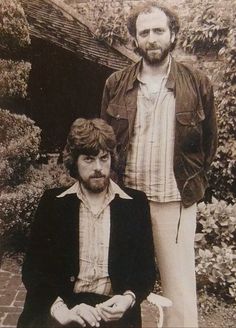 two men standing next to each other in front of a brick wall and tree covered building