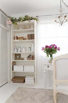 a white book shelf filled with lots of books next to a chair and table in front of a window