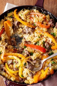 a skillet filled with pasta and vegetables on top of a wooden table next to a napkin