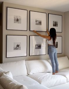 a woman hanging pictures on the wall above a white couch in front of a living room