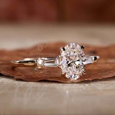 an oval diamond engagement ring sitting on top of a piece of wood next to a leaf