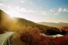 the sun shines brightly on a winding road near a lake and mountains in the distance