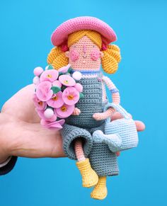 a hand holding a crocheted doll with flowers in it's lap and wearing a pink hat