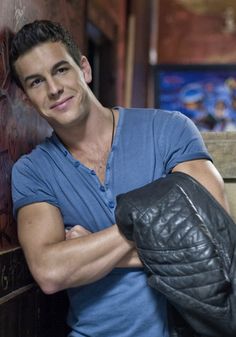 a man with his arms crossed leaning against a wooden wall wearing a black leather glove