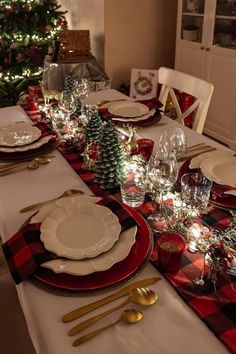 the table is set for christmas dinner with red and white plates, silverware, and evergreen trees