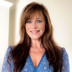 a woman with long brown hair wearing a blue shirt and smiling at the camera while standing in front of a white wall