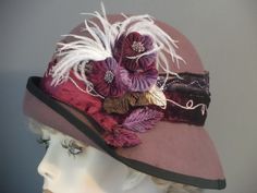 "This luscious mauve felt has been hand made using a vintage hat block. All hand made. Added flowers, feathers and 2 tone velvet band make this hat quite a statement piece. The 2.5\" band that encircles the crown is decorated with gold & lurex swirled trim, & sequins, against mauve and burgundy velvet. White small ostrich feathers, and light and dark purple flowers with wine, purple and green velvet leaves adorn the side in a lush melange. Black grosgrain ribbon trims the edge of brim. A special occasion hat for weddings, formal events, New Years, and any outdoor event. Fit 22.5\". I can adjust to fit other sizes." Handmade Fitted Vintage Fascinator, Handmade Formal Hats, Handmade Fitted Hats For Formal Occasions, Handmade Fitted Formal Hats, Elegant Brown Top Hat For Winter, Elegant Burgundy Hat For Winter, Formal Fitted Handmade Hats, Fitted Bohemian Party Mini Hats, Vintage Felt Hat For Party In Fall