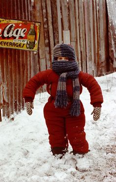 a small child in red snowsuit and hat walking through the snow with an old coca cola sign behind him