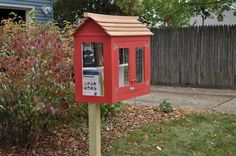 a red mailbox sitting on the side of a road next to a tree and bushes