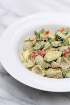 a white bowl filled with pasta and veggies on top of a marble table