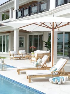 an outdoor pool area with lounge chairs and umbrellas next to the swimming pool in front of a large white house