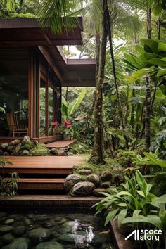 a house in the woods with plants and rocks on the ground next to it's water