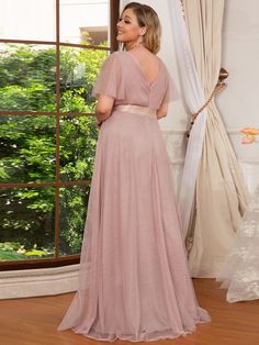 a woman standing in front of a window wearing a long pink dress with short sleeves