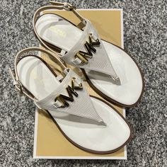 Gold-Tone Hardware Metal Logo Buckle Closure Textured Patent Leather Strap Leather Insole White T-strap Sandals With Removable Insole For Summer, White Flat Heel T-strap Sandals, White Flip Flops With Single Toe Strap, Chic White T-strap Sandals With Round Toe, White Toe Post Sandals With Cushioned Footbed, White Synthetic T-strap Sandals For Vacation, White T-strap Sandals For Beach, White Toe Post T-strap Sandals For Vacation, White Synthetic Flip Flops With Single Toe Strap