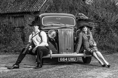 two people sitting on the front of an old fashioned car, posing for a black and white photo