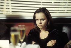 a woman sitting at a table with a glass of wine in front of her and another person behind her