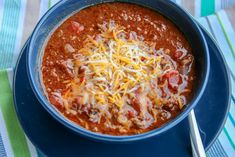 a blue bowl filled with chili and cheese on top of a striped table cloth next to a spoon