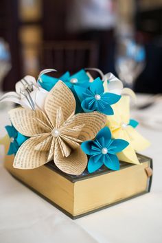 an open book with paper flowers on it sitting on top of a white table cloth