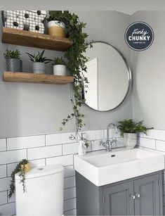 a white toilet sitting next to a bathroom sink under a round mirror and wooden shelves