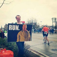 a man holding up a run sign on the side of the road with people running in the background