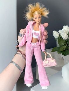 a hand holding a barbie doll wearing pink pants and a white shirt with blonde hair