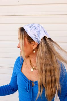 a woman with long blonde hair wearing a blue top and gold earrings, standing in front of a white wall