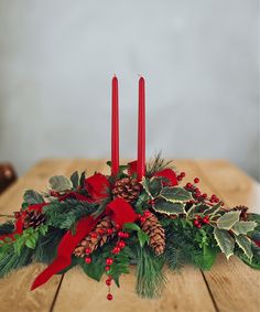 two red candles sitting on top of a wooden table next to holly and pine cones