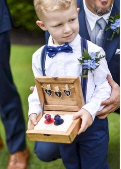 a little boy in a suit holding an open box with some jewelry on it and another man standing behind him