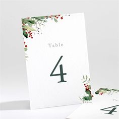 a table number card with holly leaves and berries on it, sitting next to two folded cards