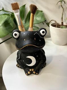 a ceramic frog with two brushes in its mouth sitting on a table next to a potted plant